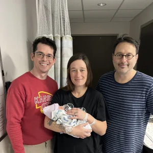 Shelby, a surrogate with Pathways To Parenthood, standing in a hospital room holding a newborn baby alongside the intended parents shortly after birth.